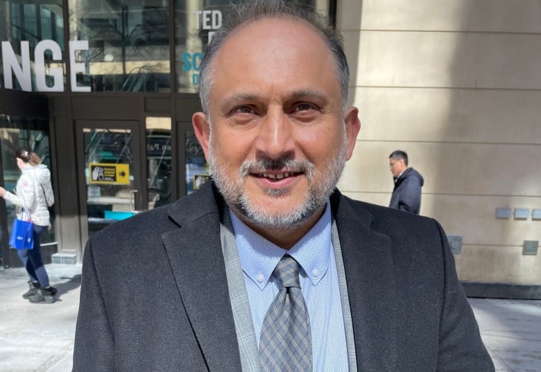 A man with grey hair and a grey beard, wearing a blue overcoat and tie, stands outside a building.