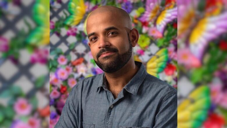 Lobo, a bearded man with a shaved head, looks into the camera in front of a colourful background of butterflies and flowers.