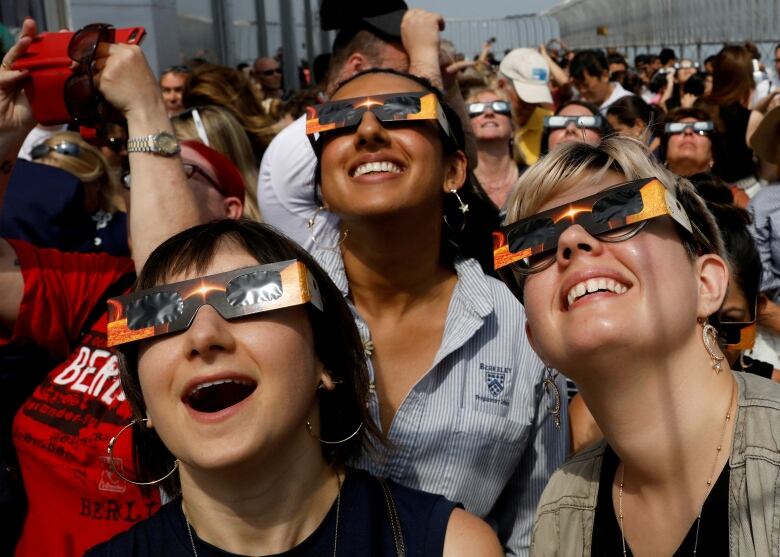 Three young women smiling, wearing eclipse glasses while looking skyward.