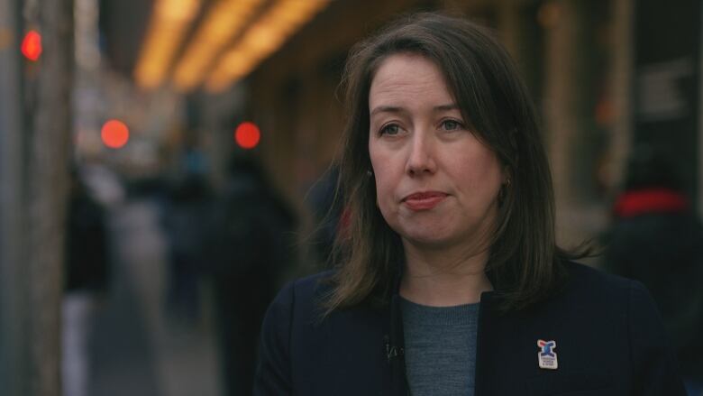 A woman with brown hair and a blazer stands on a sidewalk in Toronto.