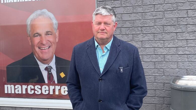 A man wearing a jacket stands outside in front of a grey brick building and a poster.