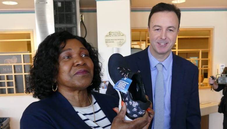 A woman is pictured holding up a black shoe to the camera as a man stands next to her, smiling.