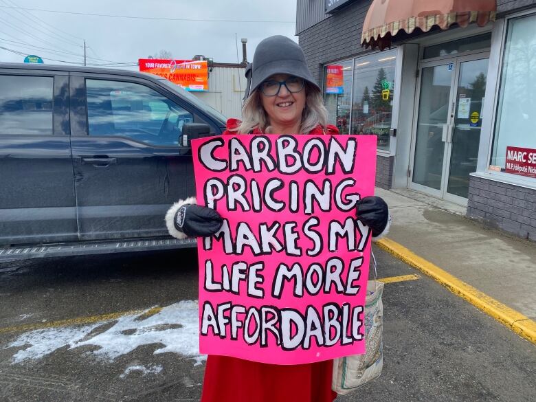 A woman wearing a hat and glasses holds a pink and white poster in a parking lot.