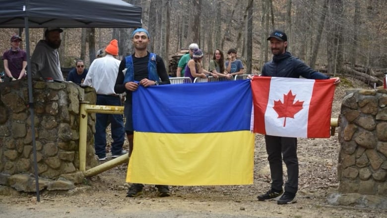 A man in dark clothes and a blue vest is holding up one side of a Ukrainian flag. On the other side is a man in dark clothing, who is also holding up a smaller Canadian flag. 