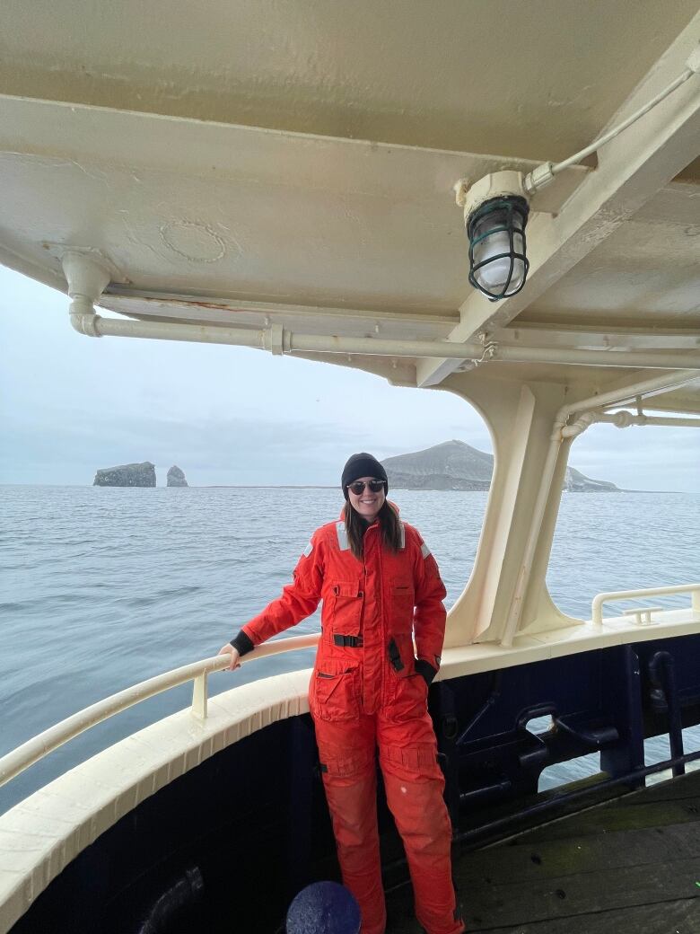 A person dressed in bright orange wearing sunglasses and looking at the camera from the edge of a boat with a backdrop of water and possibly hills in the background.