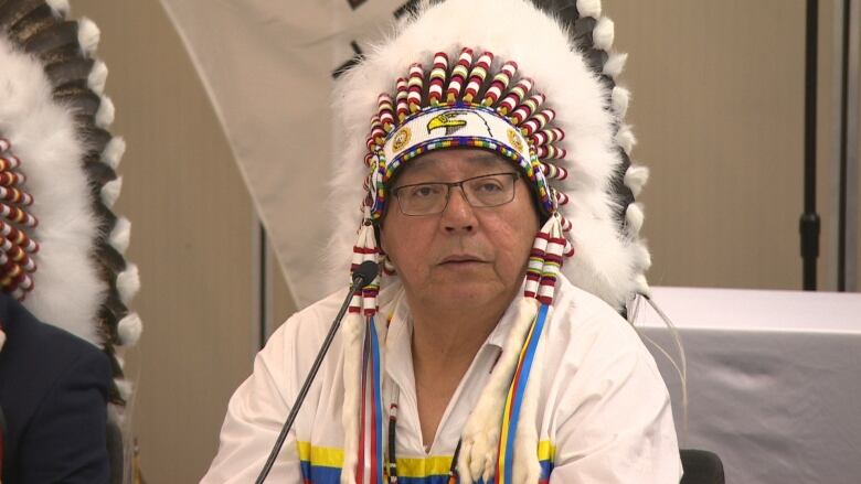 A man wearing a headdress sits behind a table in front of a microphone.