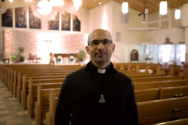 A priest stands in front of rows of pews inside a Catholic Church. He is bald and wearing glasses.