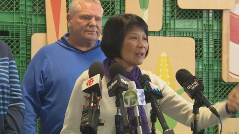 A woman speaks at a podium in front of microphones. A middle aged man stands behind her