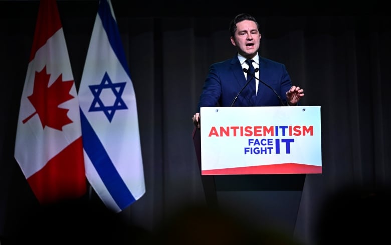 A man in a blue suit speaks at a podium, with the flags of Canada and Israel nearby.