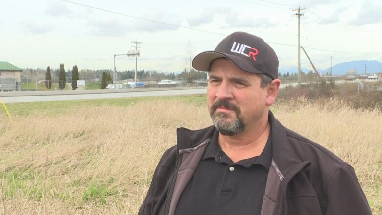 A white man wearing a baseball cap looks sullen in front of a road.