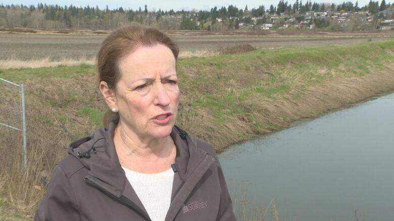 A white woman wearing a 'City of Surrey' sweatshirt speaks in front of a water body.