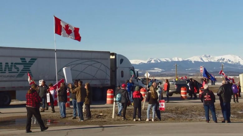 a group of people standing on a highway. a semi truck drives behind them in the distance. one person holds a Canadian flag. mountains in the back.