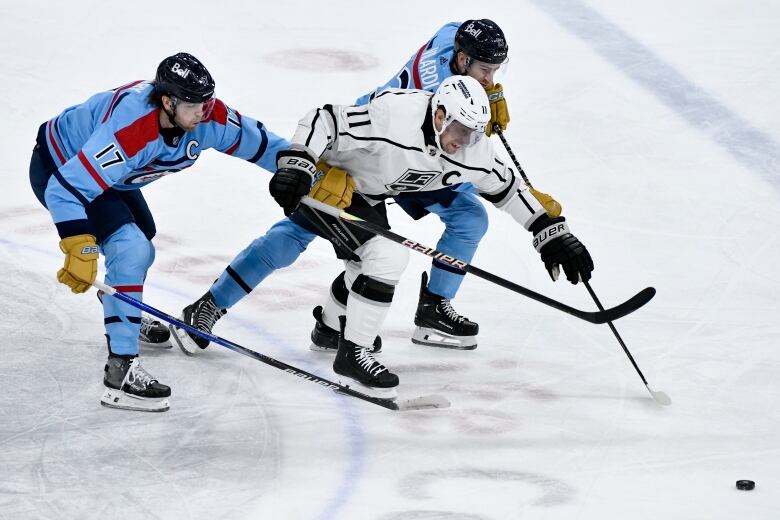 Two hockey players in blue jerseys sandwich another player of the opposite team. The puck is seen on the ice just out of reach in front of them.