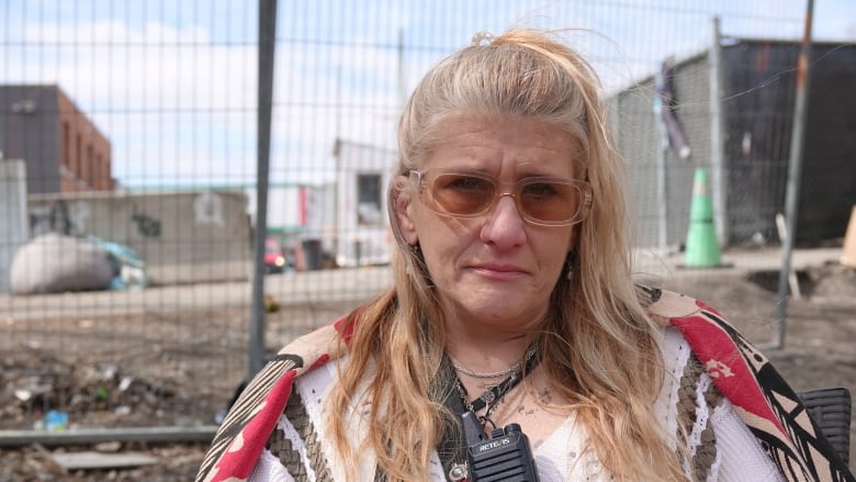 A woman with blonde hair in a ponytail stares at the camera. She's wearing sunglasses and has a walkie talkie hanging from a lanyard around her neck.