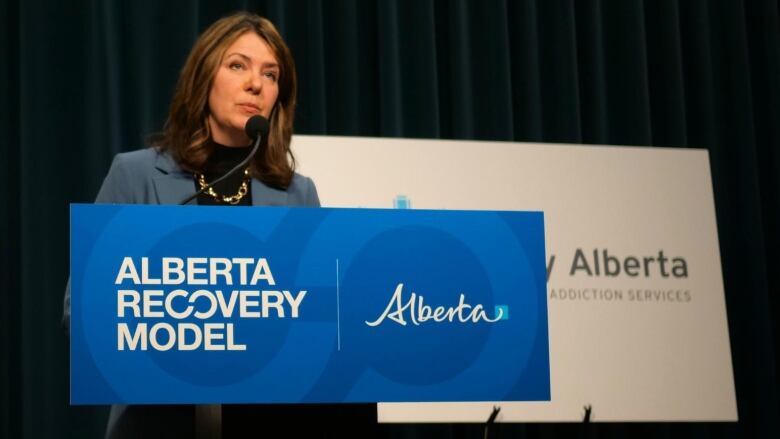 A woman with brown hair stands behind a blue podium in front of a black backdrop.