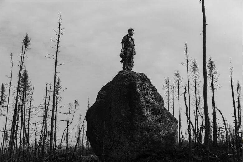 A wildfire fighter stands on a tall boulder amid charred trees.