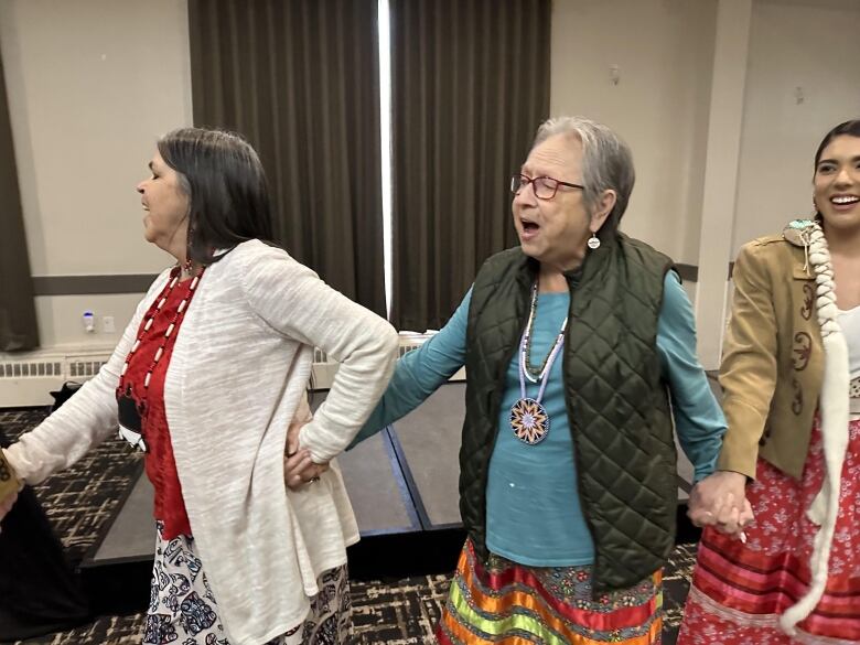 Three smiling women hold hands in a line dance.