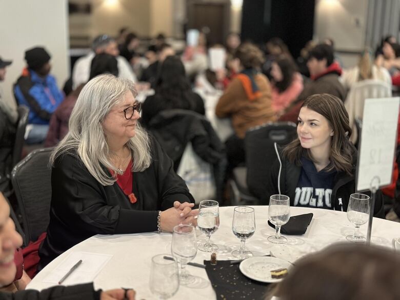 A bespectacled woman with long, white hair sits a table, smiling at a young girl with brown hair sitting next to her.