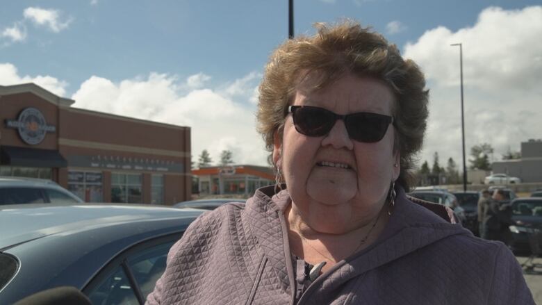 Woman with short hair and sunglasses looks into the camera while standing in a outdoor parking lot.