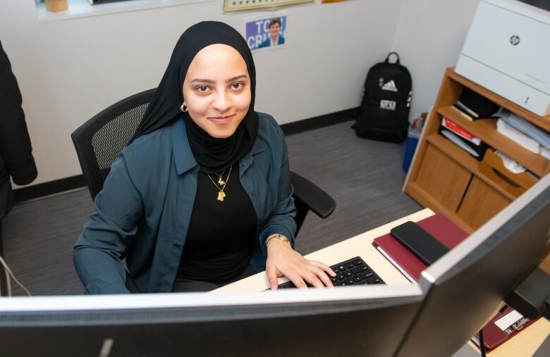 A young woman wearing a black head scarf smiles up at the camera while sitting at a desk and working on a computer.