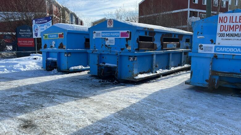 Three big bins in a lot with apartment buildings in the background. They have signs that say 'plastic,' 'mixed paper' and 'cardboard' with further instructions too small to be read.