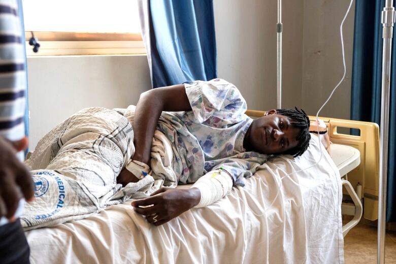 A man with short braided hair lies in a hospital bed on his side, facing the camera and clutching his mid-section. He has bandages around one arm and is attached to an IV. 