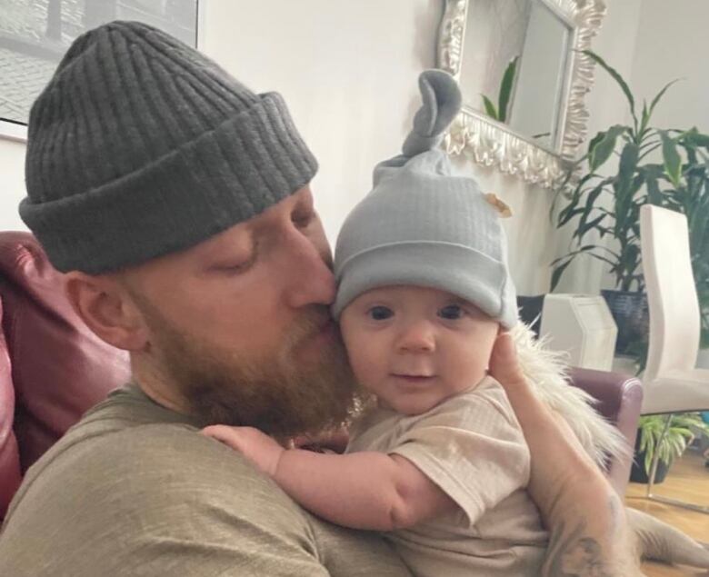 A man with tattoos and a tuque holds a baby and kisses his cheek