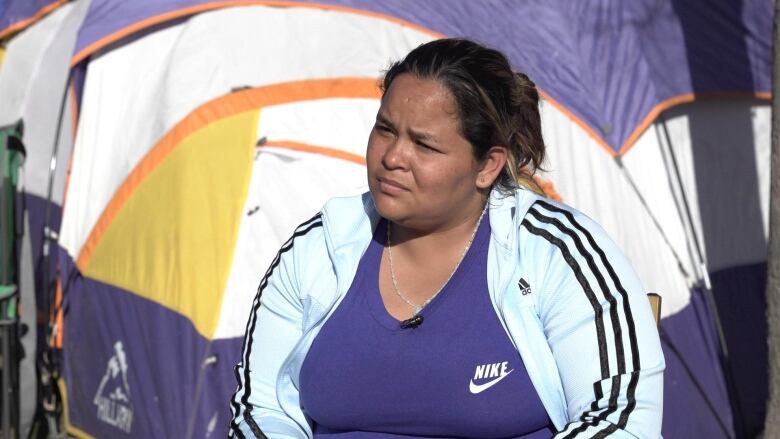 A woman sits in front of a tent. She has her hair in a bun and is wearing a light blue Adidas striped jacket and dark blue Nike shirt. 