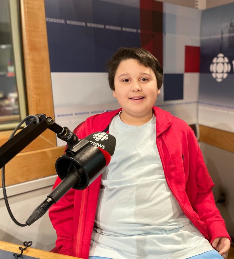 A boy with brown hair and a red button up layered over a white t-shirt sits in a radio studio, microphone in front of him.