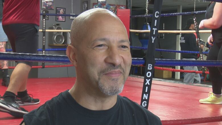 A bald bearded man smiles in front of a boxing ring.