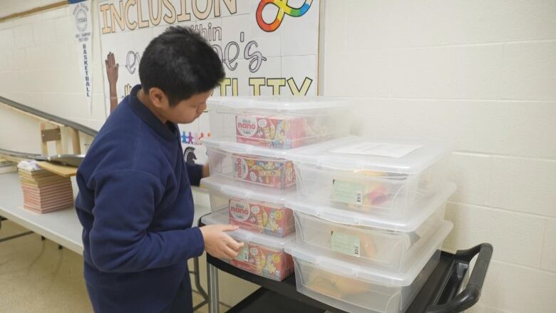 Teen boy looks at boxes filled of snacks on a rolling cart in a school building. 