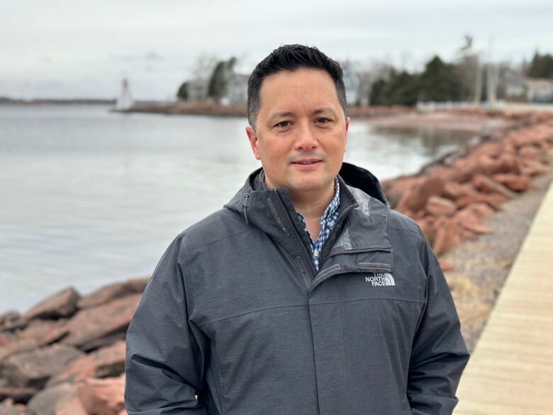 A man stands on a boardwalk with a shoreline behind him 