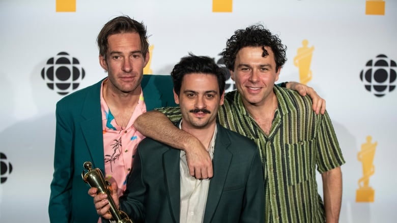 Three men stand with their arms around each other, with one holding a trophy. 