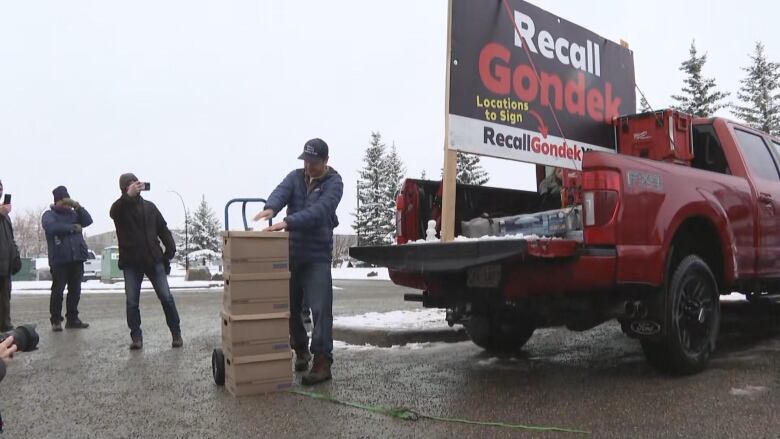 a man stands outside behind a red truck. snow is falling. other people stand behind him. he looks down at some cardboard boxes.