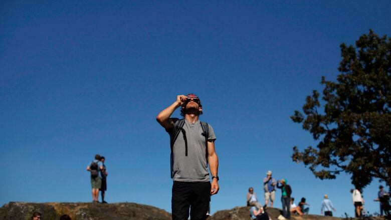 A man in a grey t-shirt and black shorts holds an eclipse viewer up to his eyes to look at a solar eclipse. The sky behind him is clear and blue.