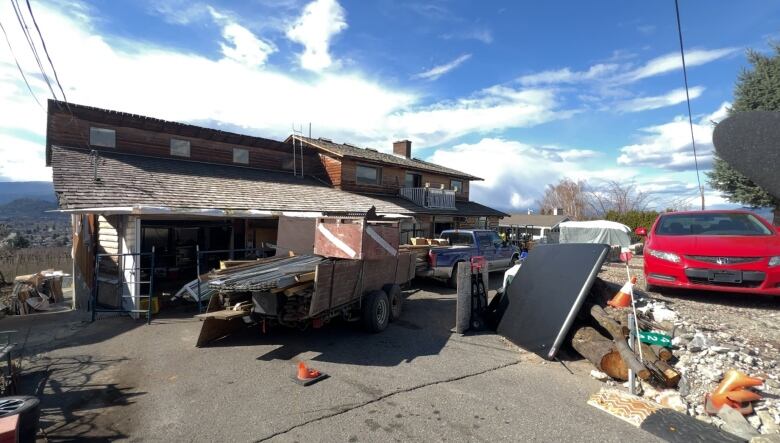 A photo of a weathered two story home on a Kelowna street with piles of building materials and a truck in front loaded with construction equipment. 