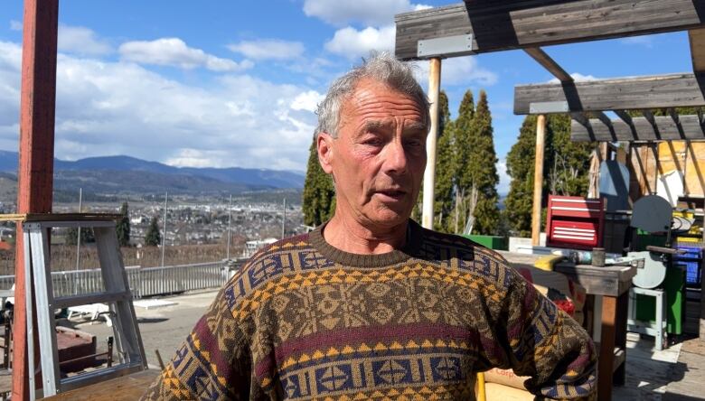 A 73-year-old man stands in his back yard on a cloudy day with hills in the distance. He is standing in a cluttered yard with a lot of construction material around him. 