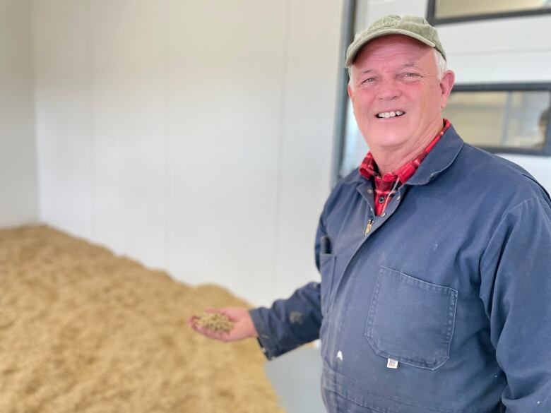 A man in blue coveralls holds a palm full of barley in his right hand. He smiles at the camera from under a green ballcap.