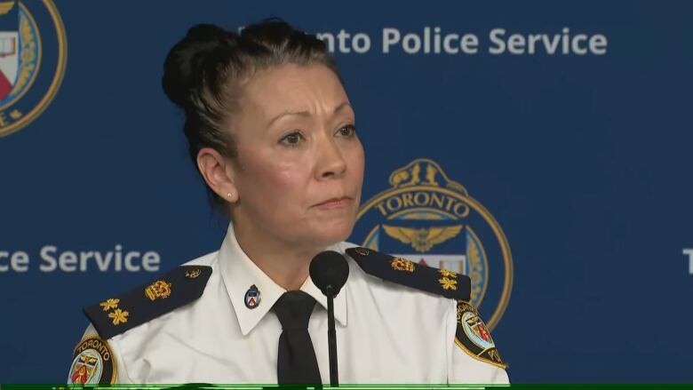 A middle-aged woman in a police uniform stands at a podium in front of a microphone. She is visible from the shoulders up.