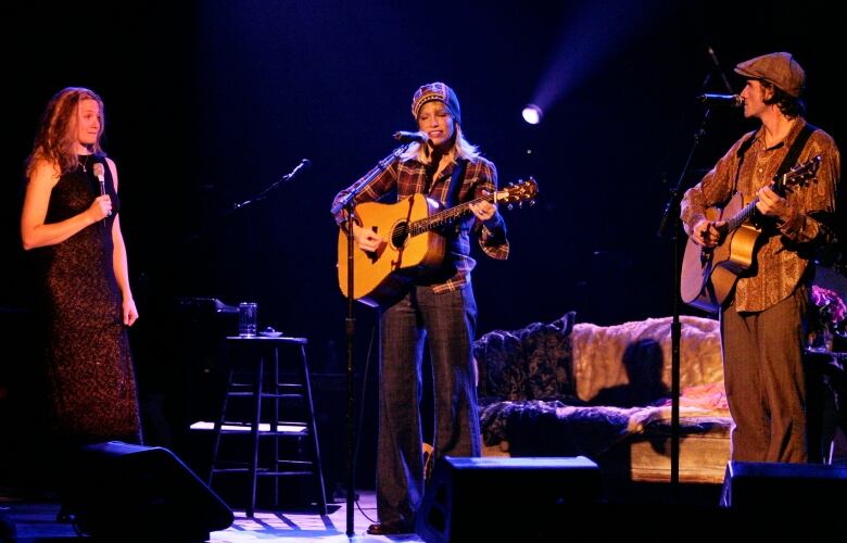 Singer Carly Simon is shown on stage with an acoustic guitar and her two adult children, one of whom is playing an acoustic guitar.