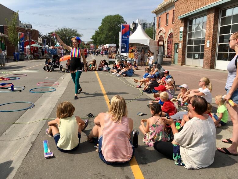 A performer in colourful clothing uses a hula hoop while children, sitting on the ground, watch.