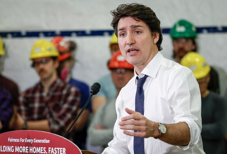 a man in a white dress shirt and blue tie speaks at a mic with people behind him