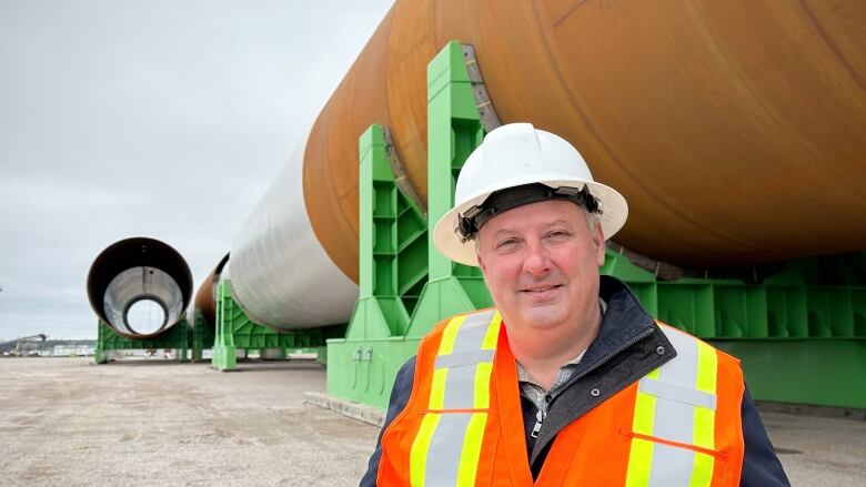 A man with a white construction helmet and an orange safety vest stands in front of a large steel tube that is lying on its side.