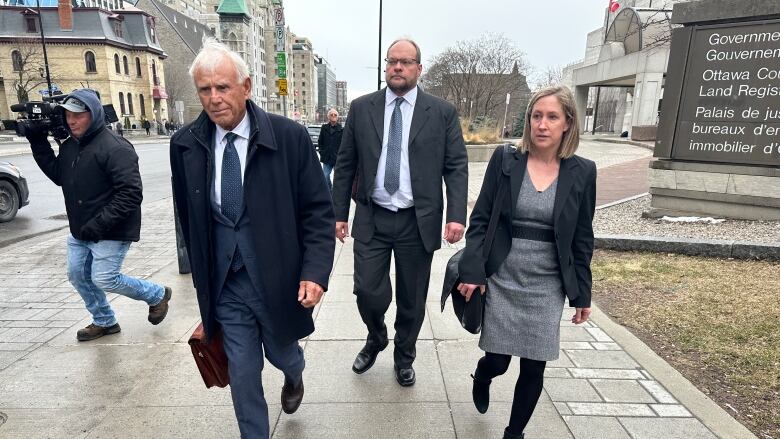 A man wearing a suit walking behind a woman and a man.