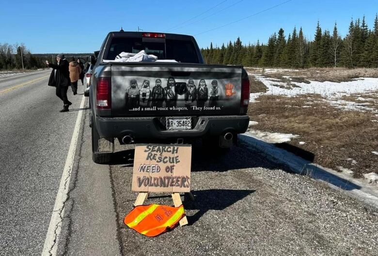 People are seen walking away from a pickup truck. An orange safety vest is seen in the foreground, on the ground near the truck.