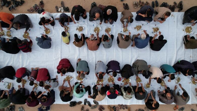 An overhead view shows people sitting in rows along on a long white sheet with plates of food in front of them