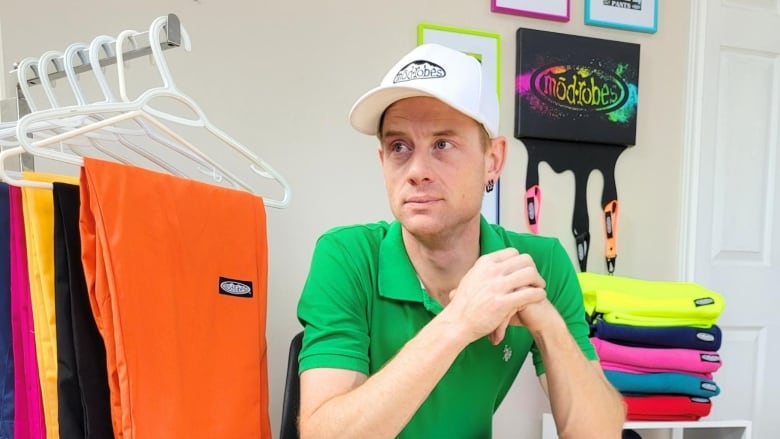 A man sits on a chair that's sitting on a checkered rug. Next to him is a rack on which is hung pairs of pants in a range of bright colours.