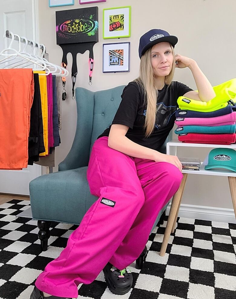 A woman in bright pink pants, a black T-shirt and ball cap sits on a chair with her elbow resting on a stack of colourful pants.