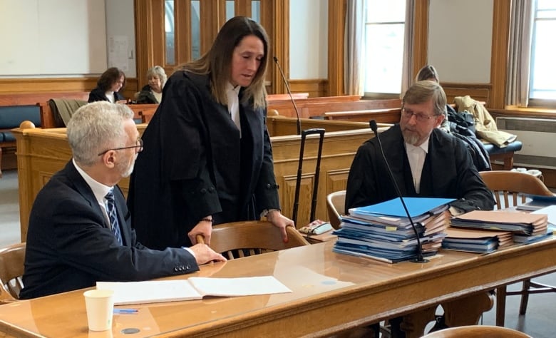 Two men sit at a wooden desk and speak with a woman standing above them. All but one of the individuals are wearing black lawyer's robes. 