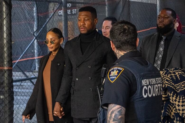 A man wearing an all-black double breasted suit walks while holding hands with a woman. They are surrounded by others wearing suits and a police officer.